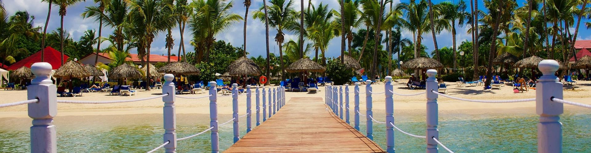 pexels-photo-247447-247447 Wooden dock leading to a tropical beach resort with palm trees and clear blue waters.